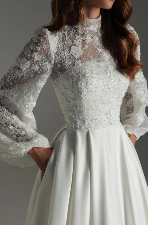 Detailaufnahme eines weißen Brautkleids mit hochgeschlossenem Spitzenoberteil, floralen Stickereien und langen transparenten Ärmeln; fließender Rock aus glattem Stoff vor neutralem Hintergrund.