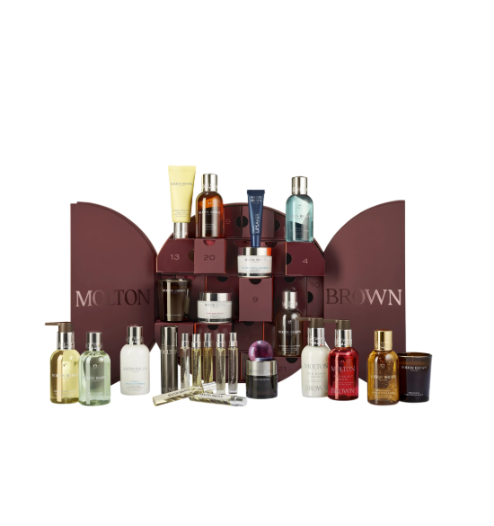 Ein geöffneter, runder, weinroter Adventskalender von Molton Brown mit kleinen, durchnummerierten Schubladen. Vor und im Kalender sind zahlreiche Pflegeprodukte der Marke, wie Duschgele, Lotionen und kleine Kerzen, drapiert.