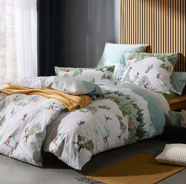 Schlafzimmer mit Holzpaneelen. Ein Bett ist mit weiß-grüner Bettwäsche mit Skifahrer- und Winterwald-Motiven bezogen, ergänzt durch eine gelbe Decke und mehrere Kissen.