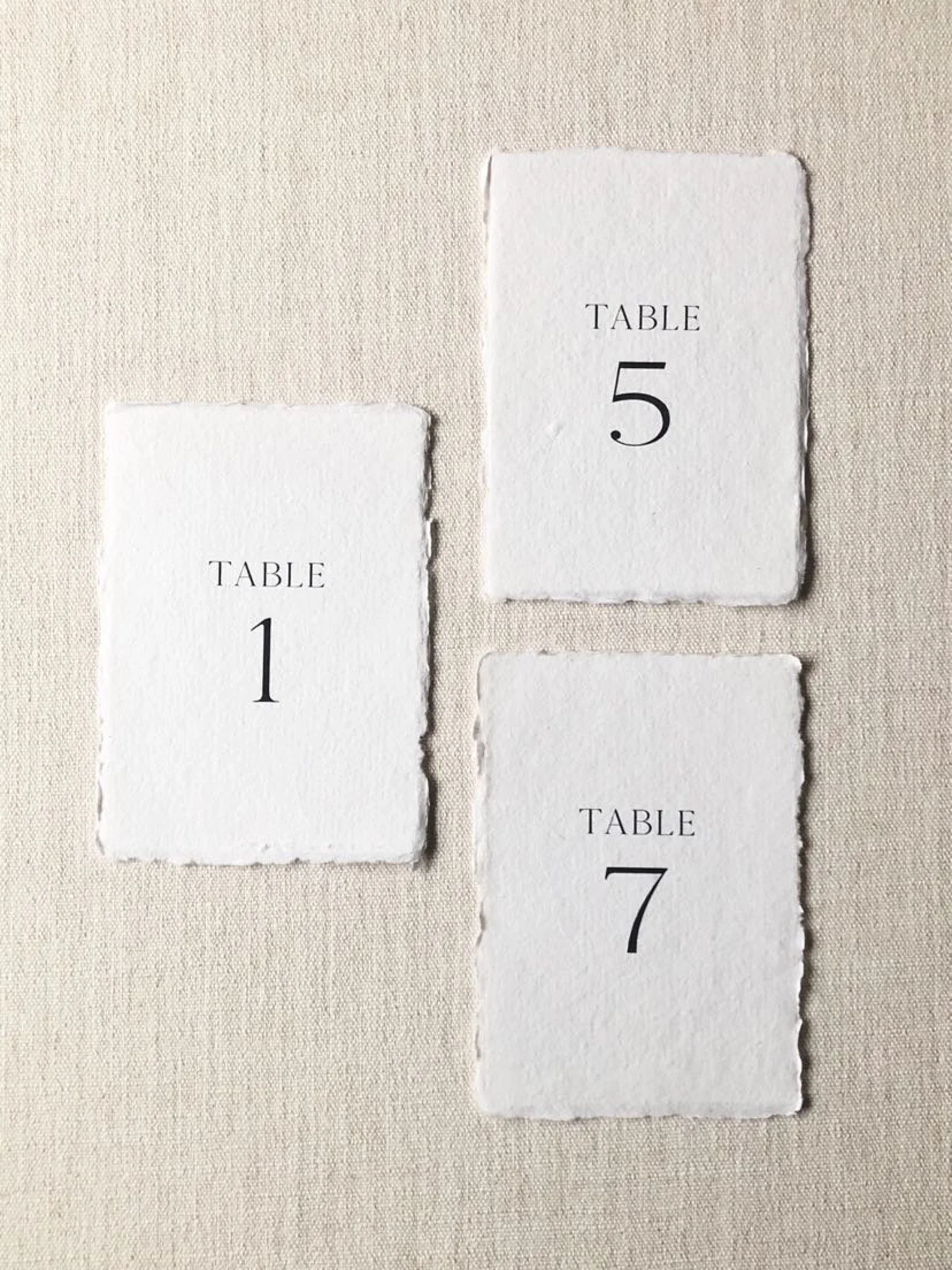 Drei hochformatige Karten aus handgeschöpftem Papier mit den schwarzen Aufdrucken „TABLE 1“, „TABLE 5“ und „TABLE 7“.