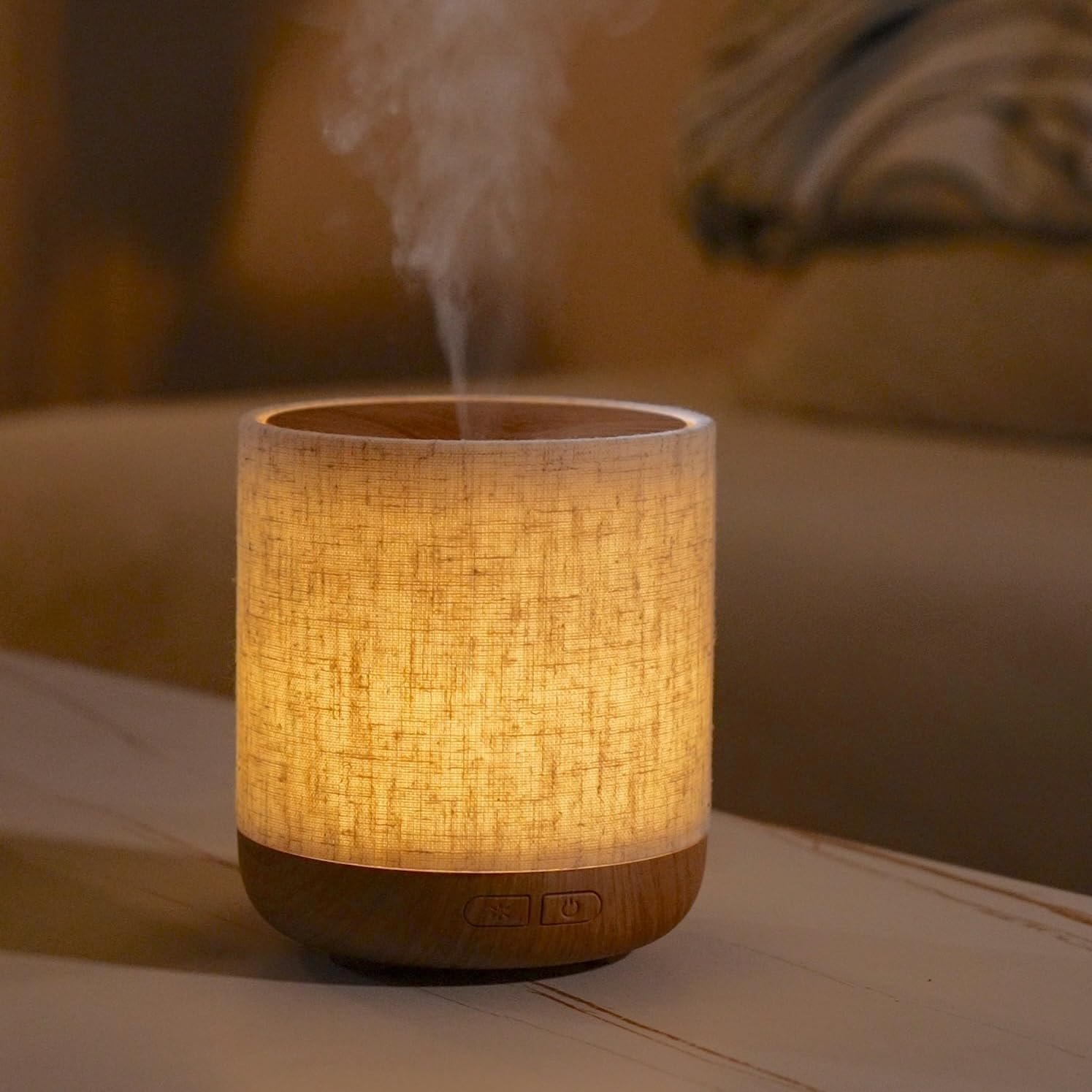 Leuchtender Aroma-Diffuser mit Holzsockel und hellem Stoffbezug auf einem Tisch. Oben steigt ein feiner Nebel aus dem Gerät auf.