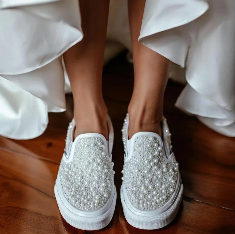 Frontale Nahaufnahme der Brautschuhe: weiße Slip-On Sneaker voll mit Perlen und kleinen Strasssteinen, im Hintergrund Saum eines fließenden weißen Kleides auf glänzendem Holzboden.