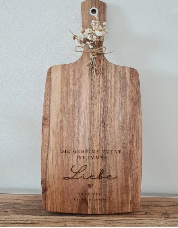 Rustikales Holz-Schneidebrett als personalisiertes Hochzeitsgeschenk mit der Gravur „Die geheime Zutat ist immer Liebe“, den Namen des Brautpaares und dem Hochzeitsdatum, verziert mit Trockenblumen am Griff.
