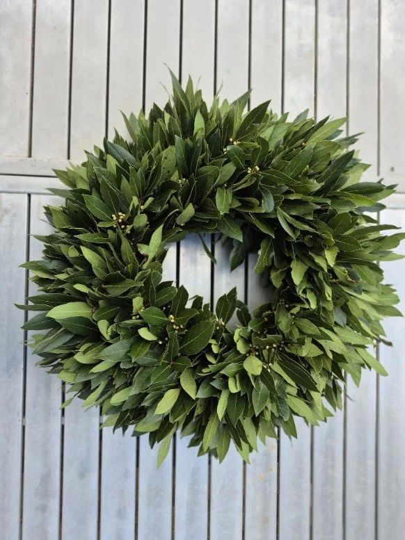 Schlichter, dichter Kranz aus frischen Lorbeerblättern mit vereinzelten gelb-grünen Knospen, vor grauen Metall- oder Holzlatten hängend.