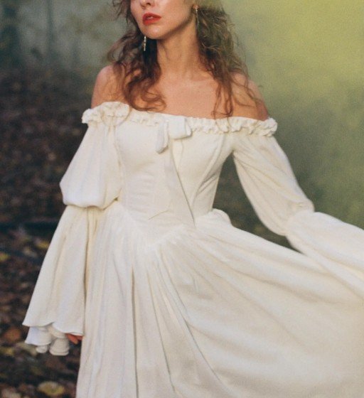 Braut in schulterfreiem Vintage-Kleid mit Korsett, gerüschtem Ausschnitt, kleiner Schleife und weiten Trompetenärmeln. Analoge Fotografie, nostalgische Stimmung, erdige Naturtöne.