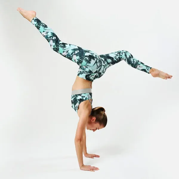 Frau im Handstand in Sport-BH und Leggings mit türkis-schwarzem Palmenmuster vor hellem Hintergrund