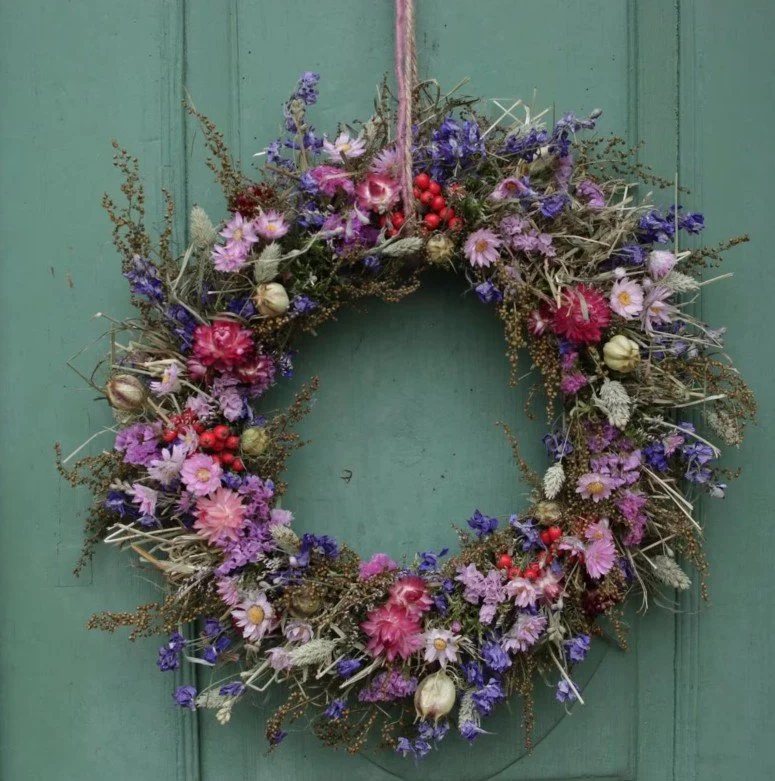 Rustikaler, wild gebundener Türkranz aus getrockneten Blumen in Lila-, Pink- und Violetttönen, kombiniert mit naturfarbenen Gräsern, beigen Mohnkapseln und roten Beeren. Hängt an Juteband an grüner Holztür.