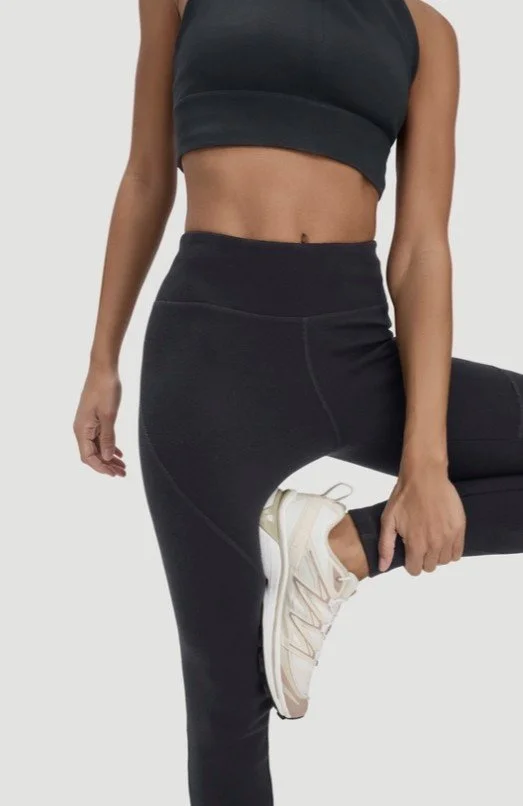 Frau in schwarzem Sportoutfit (High-Waist Leggings und passender Sport-BH) und hellen Sneakern bei einer stehenden Dehnübung vor hellem Hintergrund.