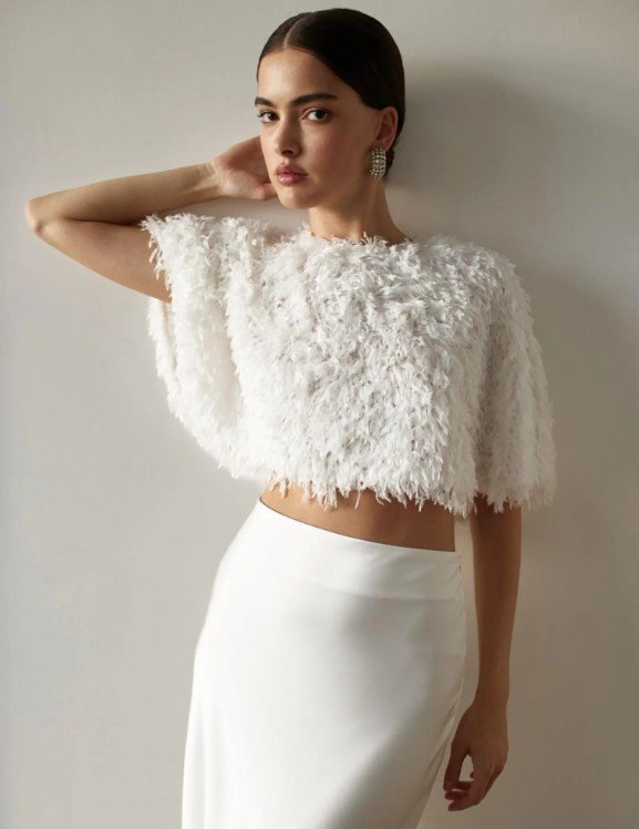 Braut trägt einen kurzen, flauschigen Pullover mit Federn-Optik über einem schlichten weißen Rock – modernes Brautoutfit mit Fashion-Statement.
