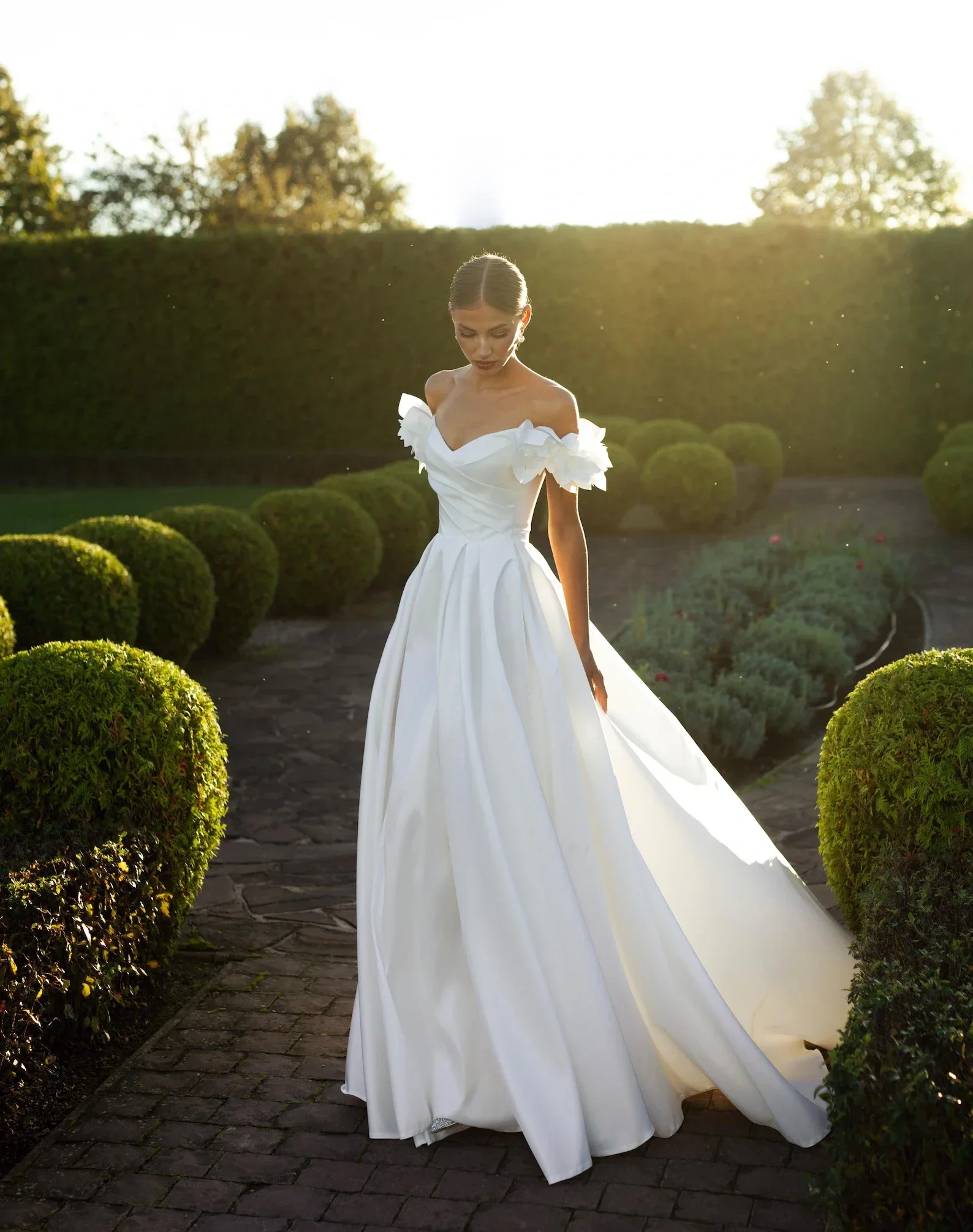 Braut in weißem Off-Shoulder-Brautkleid mit 3D-Blütendetails an den Schultern und weitem A-Linien-Rock in einem eleganten Garten bei Sonnenuntergang – handgefertigtes Brautkleid von Alma de Luz Atelier