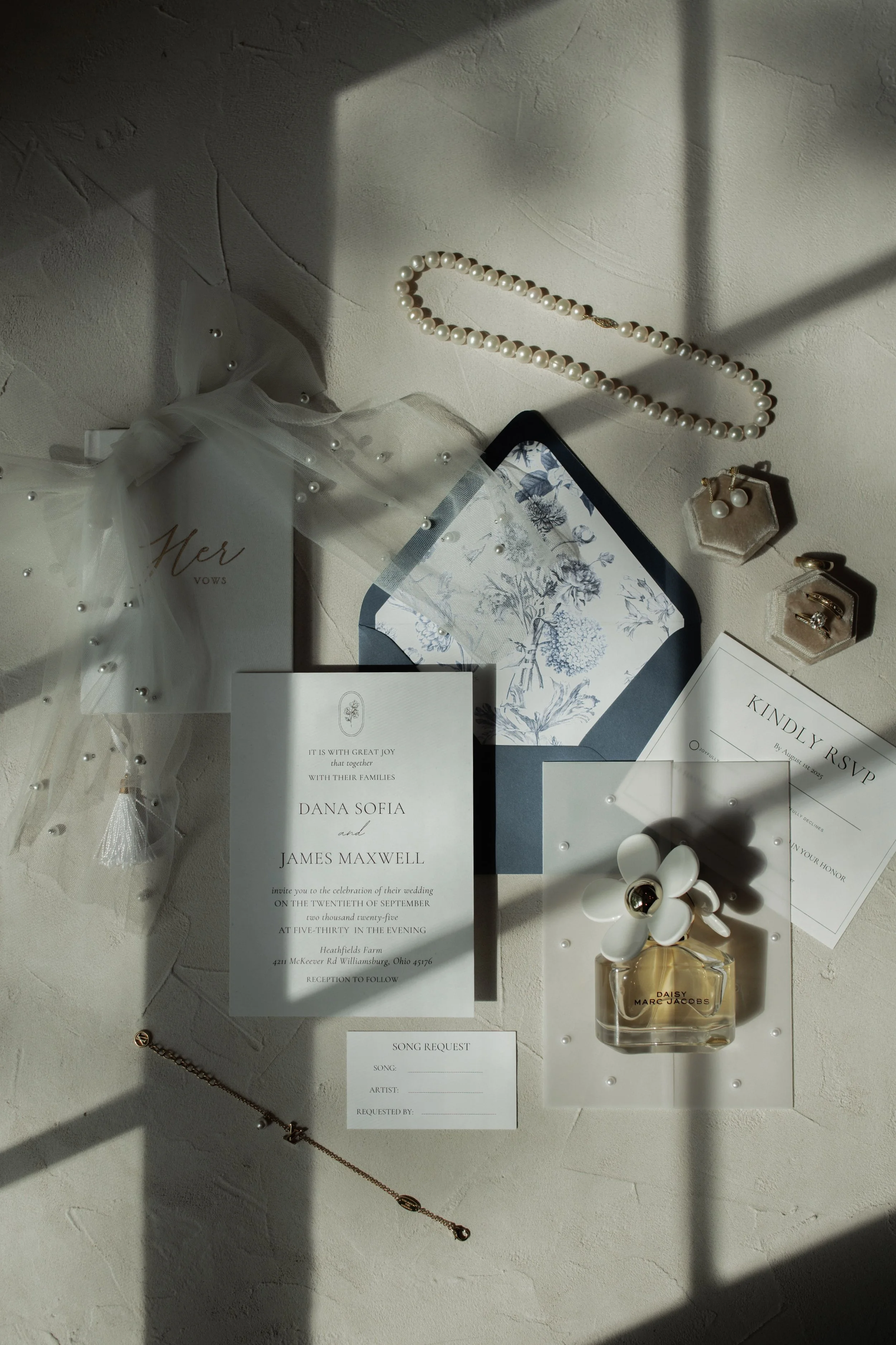 Ein Hochzeits-Einladung sowie Schmuck, Parfüm, und andere Dekorationsartikel auf einer hellen Wand. Die Einladung ist für die Hochzeit von Dana Sofia und James Maxwell am 25. September 2025.