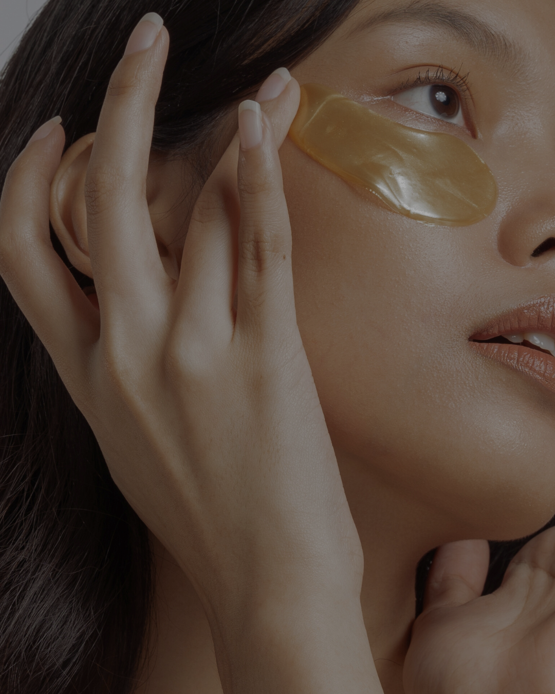 Junge Frau mit strahlender Haut legt goldene Hydrogel-Augenpads unter die Augen; ein Pad an Wange und Ohr. Gepflegte Hände mit klarem Lack, nudefarbene glänzende Lippen. Minimalistischer, neutraler Hintergrund.