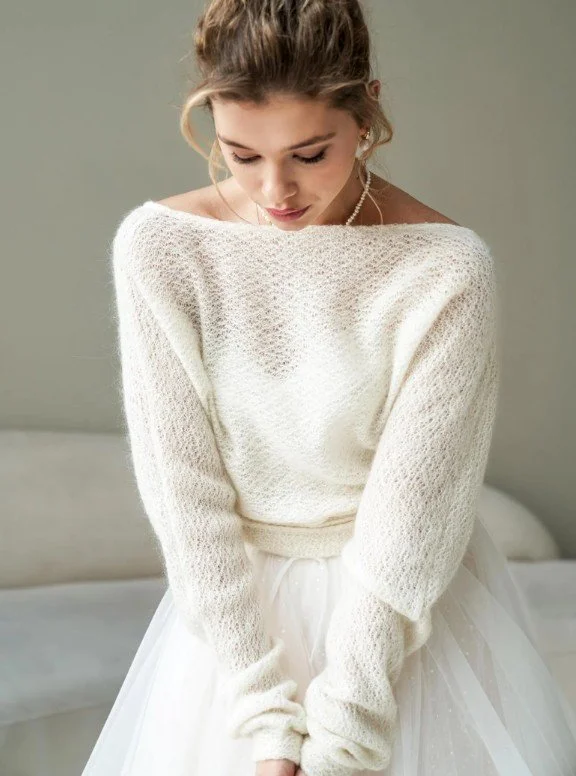 Braut in einem locker gestrickten, cremefarbenen Pullover mit weitem Ausschnitt und zartem Tüllrock, die sanft nach unten blickt.