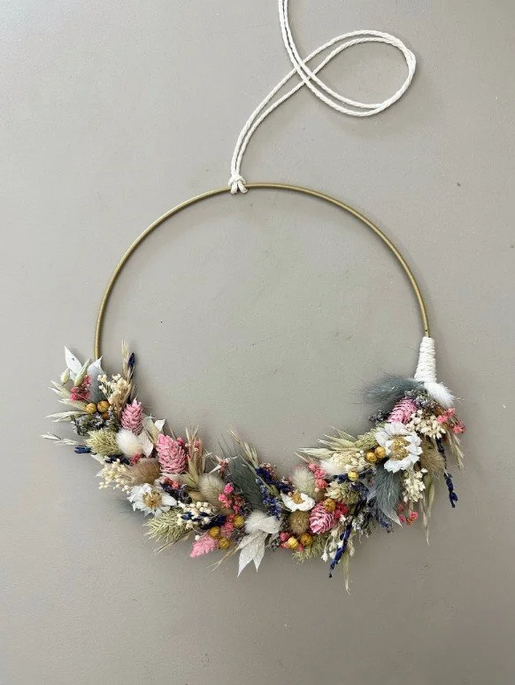 Goldener Metallring (Flower Hoop) an weißer Kordel vor grauer Wand. Unterer Halbkreis asymmetrisch mit Trockenblumen: rosa und graublaue Gräser, gebleichte weiße Blätter und kleine gelbe Blütenköpfe. Enden sauber mit weißem Garn am Ring fixiert.