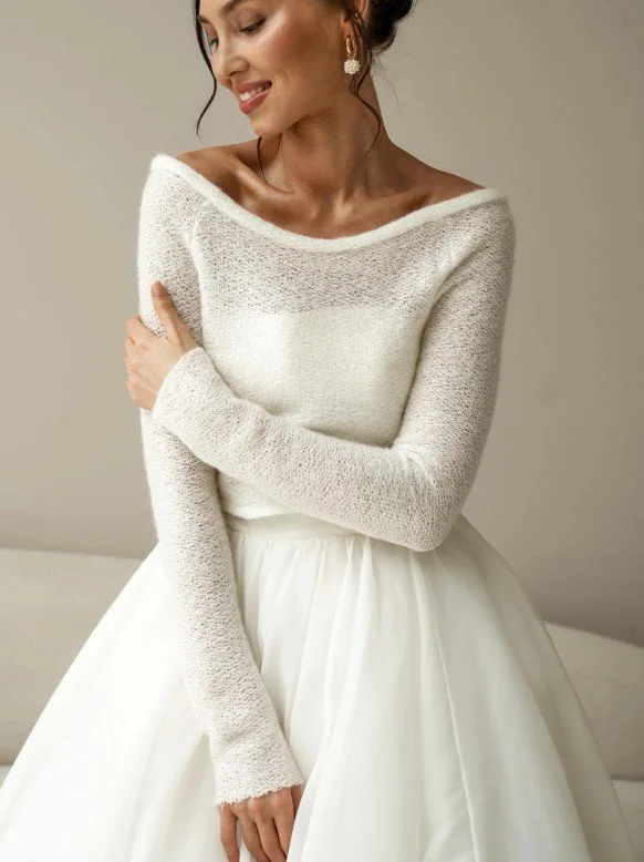 Lächelnde Braut trägt einen feinen weißen Pullover mit Carmen-Ausschnitt und voluminösem Tüllrock in einem neutralen Raum.