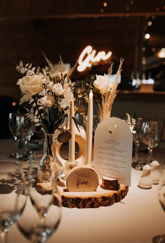 Ein atmosphärisch beleuchteter Hochzeitstisch mit einer runden Holzscheibe als Untersetzer, auf der ein Holzaufsteller mit der Aufschrift „TABLE five“ steht.