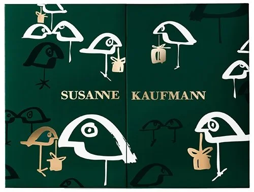 Ein dunkelgrüner Adventskalender der Marke Susanne Kaufmann. Das Cover zeigt mittig den goldenen Markenschriftzug und ist mit stilisierten Vögeln in Weiß, Schwarz und Gold verziert, von denen einige kleine Geschenke im Schnabel tragen.