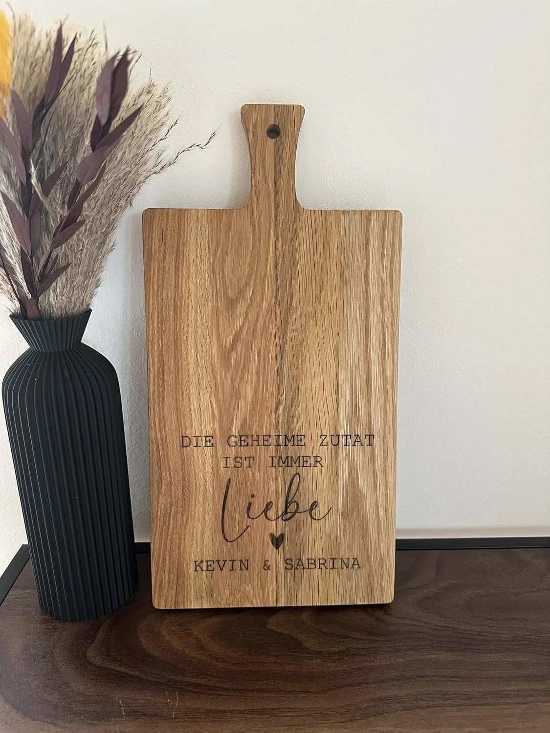 Ein hölzernes Schneidebrett mit der Gravur „DIE GEHEIME ZUTAT IST IMMER Liebe“ und den personalisierten Namen „KEVIN & SABRINA“. Es steht dekorativ neben einer schwarzen Vase mit Trockenblumen.