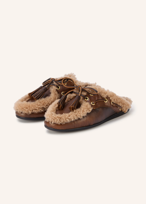 Braune Leder-Mules im Bootsschuh-Stil mit Schnürung, Quasten und einem auffälligen, hellbraunen Lammfell-Besatz entlang der vorderen Naht und im Innenfutter.