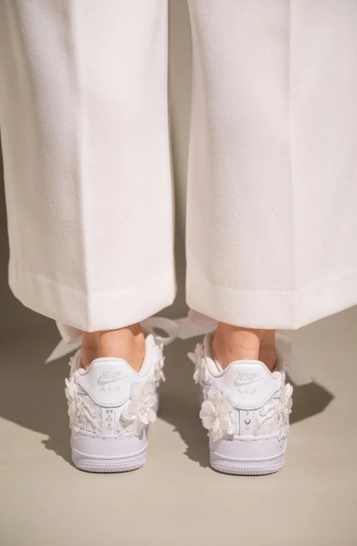 Rückansicht einer Braut in weiten weißen Hosen und personalisierten Nike Air Force 1: weiße Sneaker mit 3D-Spitzenblumen, Perlen, eingesticktem Hochzeitsdatum und Initialen, große Satin-Schleifen als Schnürsenkel.