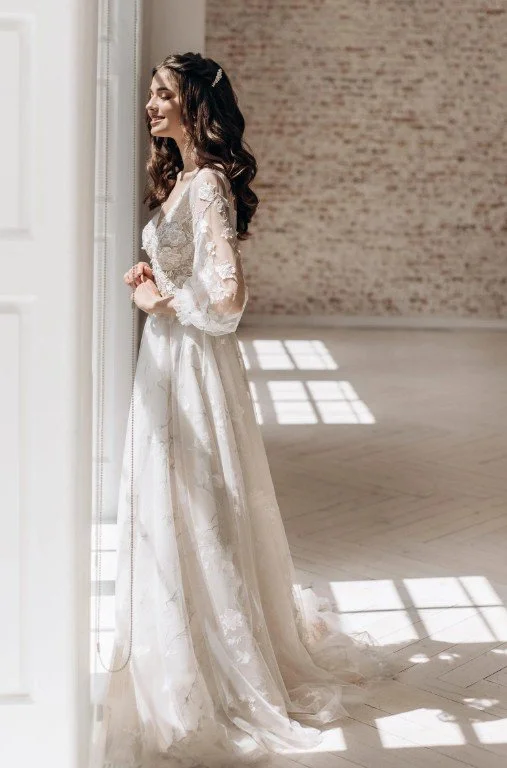 Braut am Fenster in einem langärmeligen Hochzeitskleid mit floraler Spitze und transparenten Ärmeln.