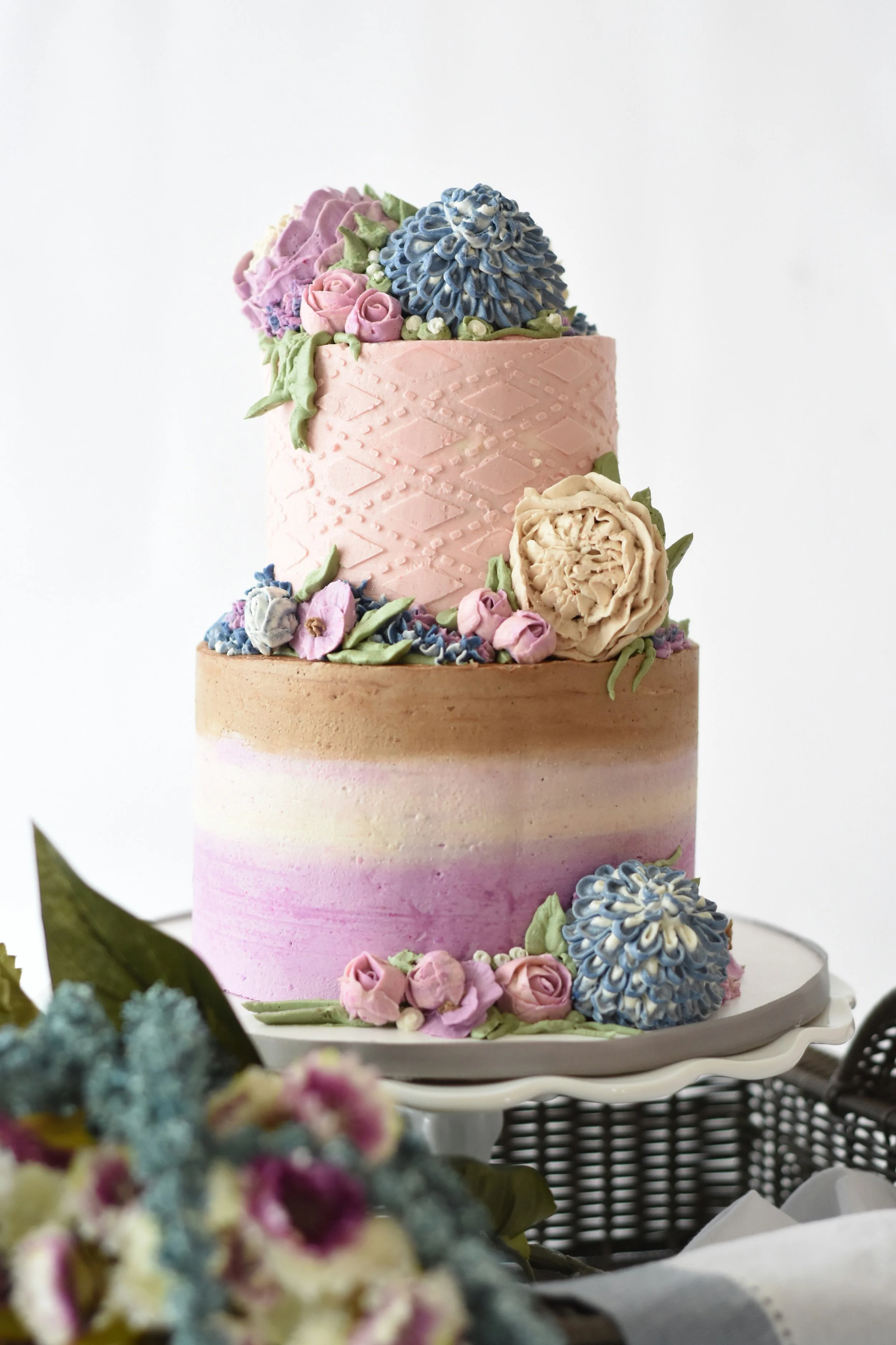 Zweistöckige Torte: untere Etage Ombré von Weiß über Beige zu kräftigem Lila/Flieder, glatt; obere Etage zartrosa mit Rautenmuster. Üppig verziert mit kunstvollen Buttercreme-/Zuckerblumen