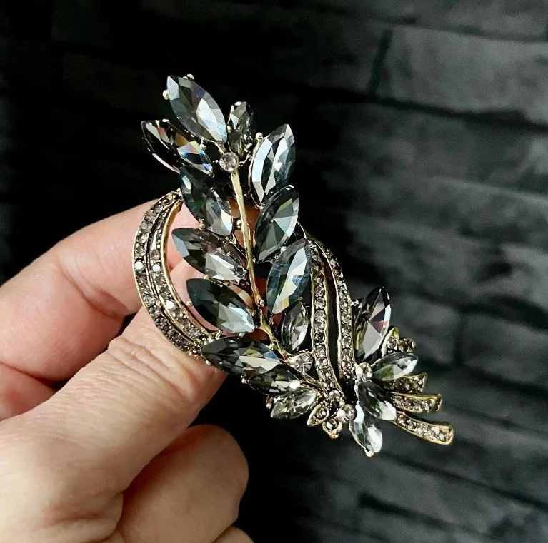 Detailaufnahme einer opulenten, blattförmigen Statement-Brosche, die mit zahlreichen grauen und klaren Kristallen in Marquise-Form besetzt ist und vor einer dunklen Wand präsentiert wird.