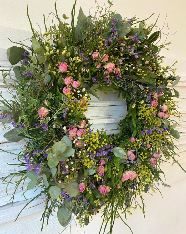 Großer, texturreicher Blumenkranz vor weißer Holzwand aus rosa Rosenknospen, gelben Blüten, lila Strandflieder und grünen Zweigen (Eukalyptus, Koniferengrün).