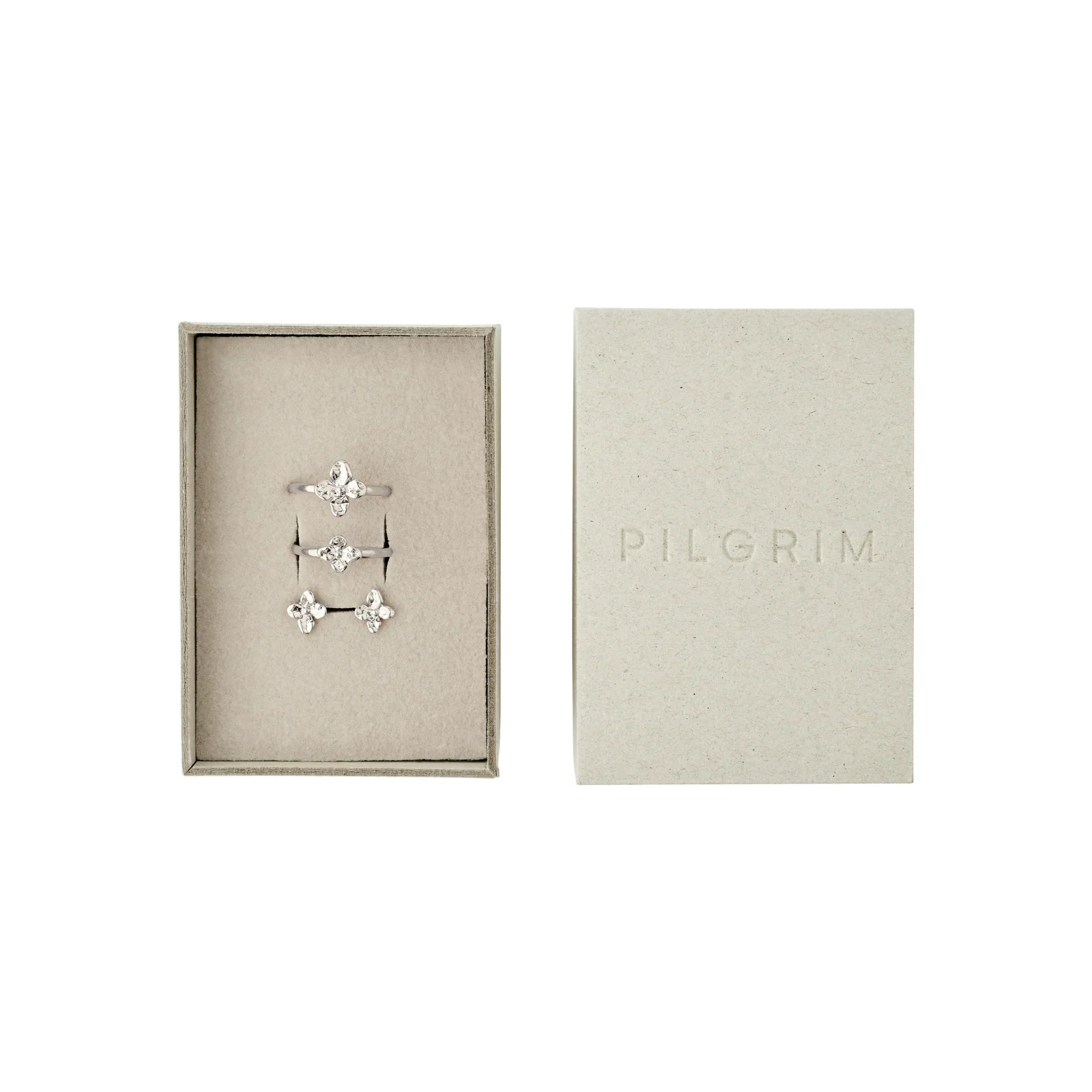 Geöffnete, hellgraue Schmuckbox von PILGRIM. Auf dem Polster liegen zwei silberne Ringe und ein Paar Ohrstecker, jeweils mit einem glitzernden Blumenmotiv.