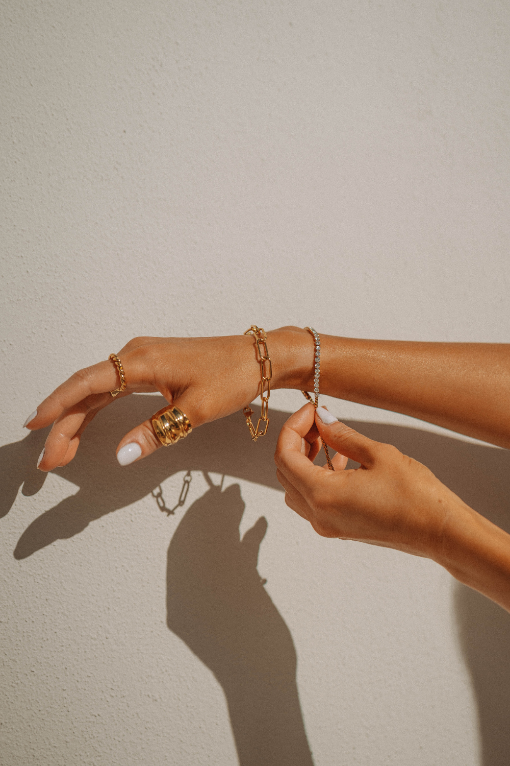 Hände mit mehreren Gold- und Silberschmuckstücken wie Ringen, Armbändern und Ketten, die gegen eine helle Wand gehalten werden.