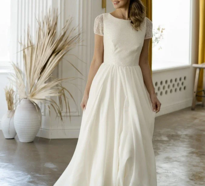 Elegantes A-Linien Brautkleid mit kurzen Spitzenärmeln, einem schlichten Mieder und einem fließenden Rock, stehend neben einer Vase mit Trockenblumen.