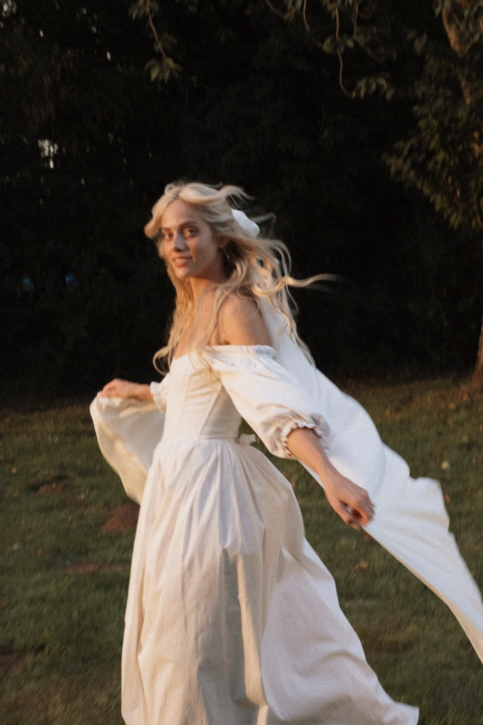 Lächelnde Braut in schulterfreiem Vintage-Kleid mit weiten Puffärmeln und weißer Schleife im lockeren Haar dreht sich auf einer Wiese. Analoge Fotoaufnahme fängt Bewegung und warmes Abendlicht im
