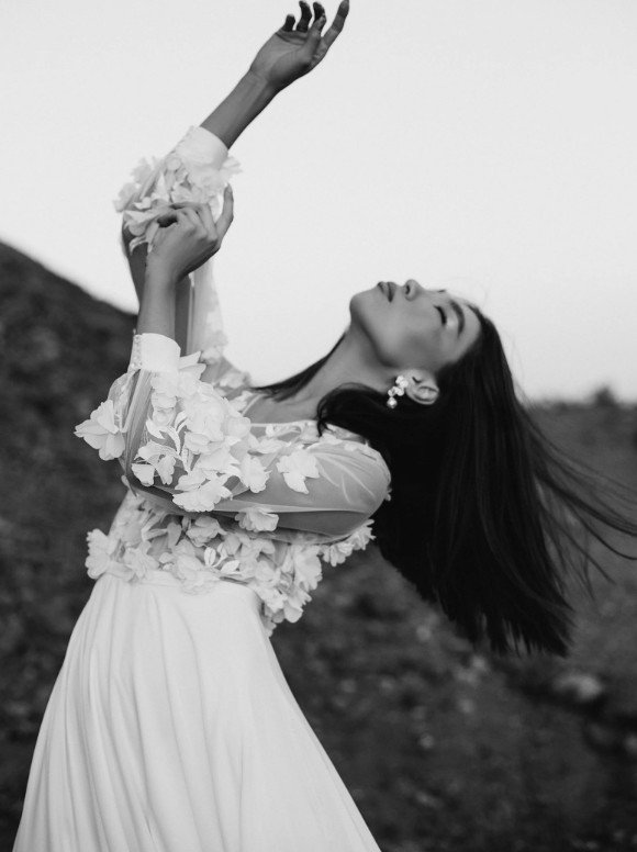 Schwarz-weiß-Fotografie einer Braut in einem dynamischen Pose unter freiem Himmel. Sie trägt ein Oberteil mit dreidimensionalen Blütenapplikationen und langen Ärmeln zu einem fließenden Rock.