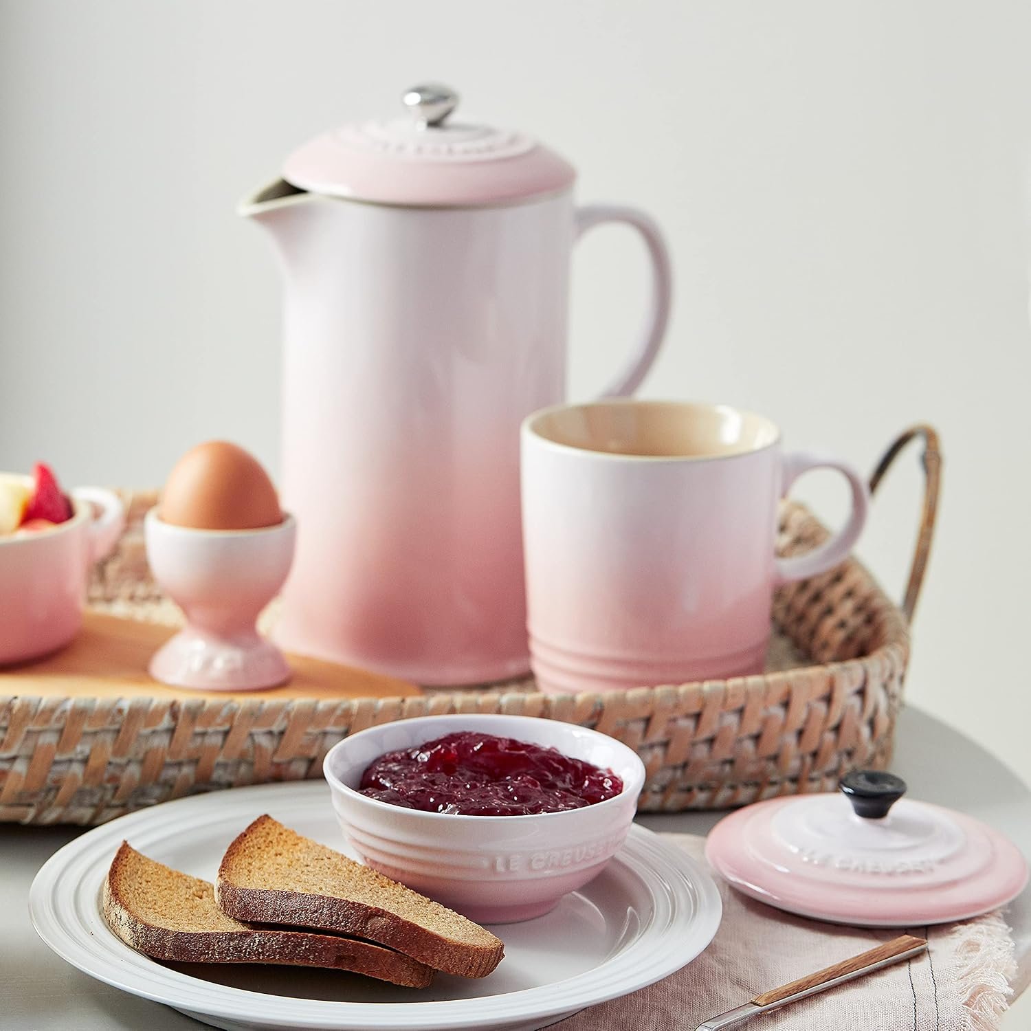 Rosa-weißes Le Creuset Frühstücksgeschirr. French Press, Becher und Eierbecher auf einem Tablett. Vorne ein Teller mit Brot und roter Marmelade im Schälchen.