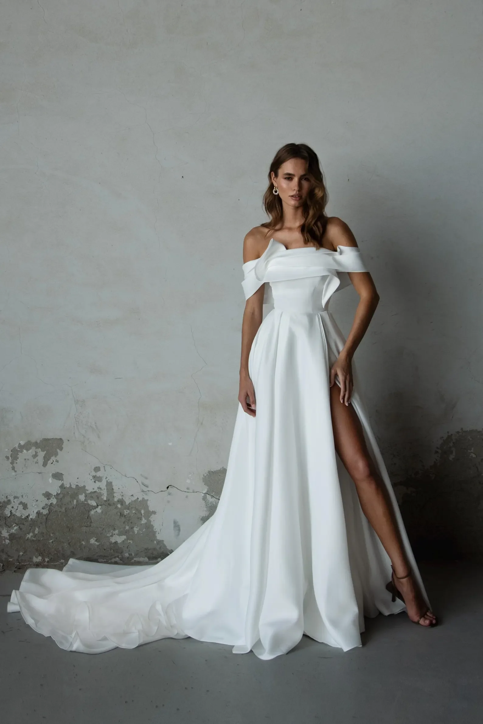Braut in minimalistischem weißem Off-Shoulder-Brautkleid mit tiefem Beinschlitz und elegantem Schleppe – modernes handgefertigtes Brautkleid von Alma de Luz Atelier