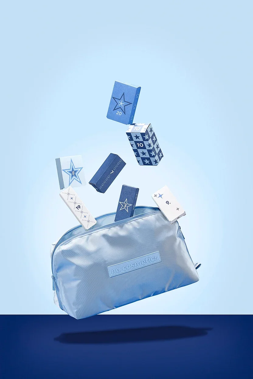 Ein Adventskalender der Marke nø cosmetics. Das Motiv zeigt eine geöffnete, hellblaue Kosmetiktasche, aus der mehrere kleine, nummerierte Schächtelchen in verschiedenen Blau- und Weißtönen mit Sternenverzierung herauszufliegen scheinen.