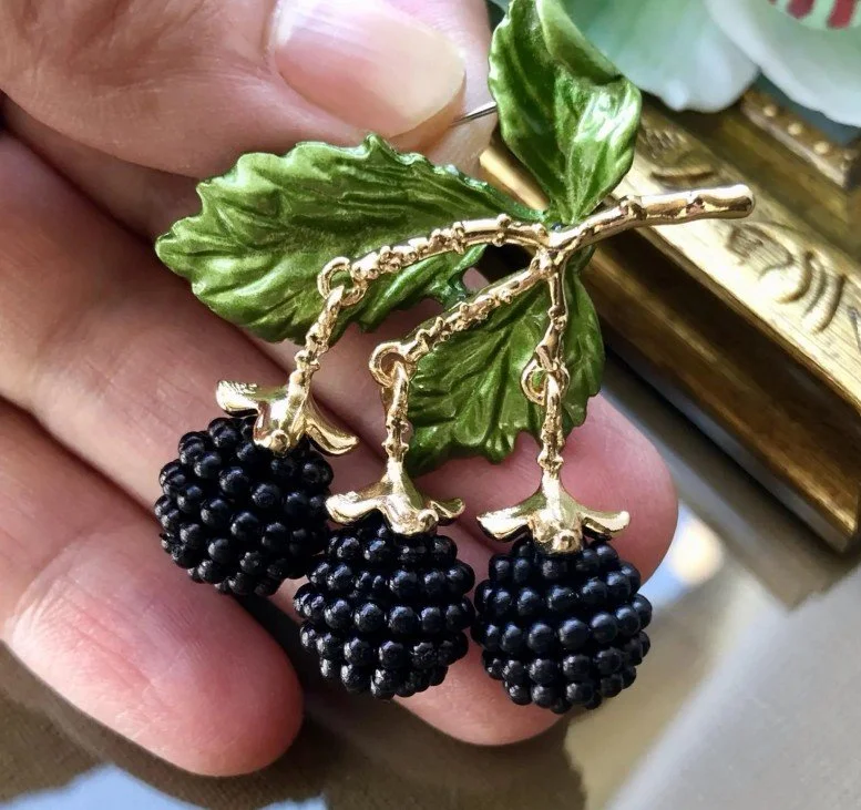 Nahaufnahme einer hochwertigen Brosche in Form von drei schwarzen Brombeeren, die an goldenen Stielen unter zwei grünen Emaille-Blättern hängen. Die Brosche wird zwischen zwei Fingern gehalten. Dekorativer Broschen Schmuck im Vintage-Stil.