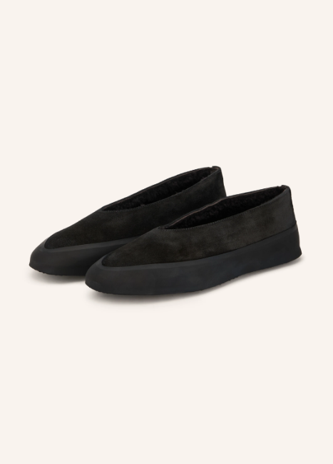 Ein Paar schwarze, flache Schlupfschuhe aus mattem Wildleder mit einer dicken, glatten Gummisohle und hochgezogener Zehenkappe.