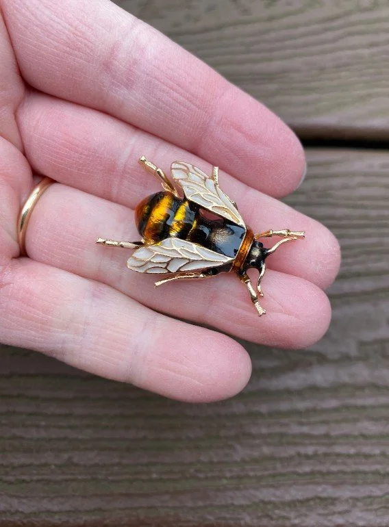 Detailaufnahme einer goldenen Bienen-Brosche mit gelb-schwarzer Emaille-Füllung und fein ausgearbeiteten, weiß-goldenen Flügeln, die flach auf einer Handfläche präsentiert wird.