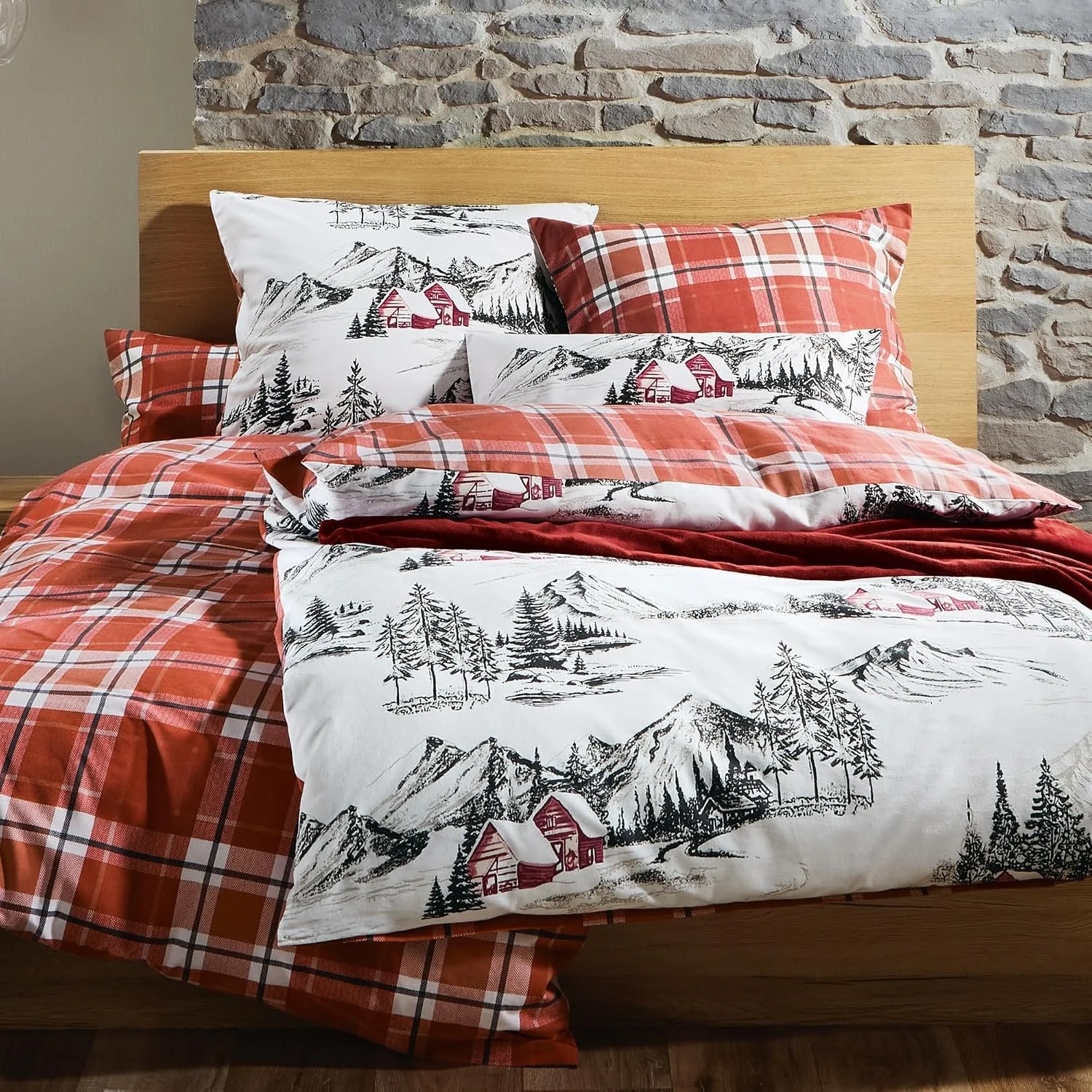 Holzbett mit rot-weiß karierter Bettwäsche und winterlichen Gebirgs- und Hüttenmotiven vor einer rustikalen Natursteinwand. Auf dem Bett liegen mehrere Kissen und eine dunkelrote Kuscheldecke.