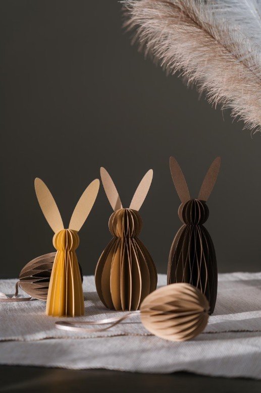 Moderne Osterdeko: Drei minimalistische Papier-Hasen (Wabenbälle) in Senfgelb und Brauntönen auf einem Tisch.