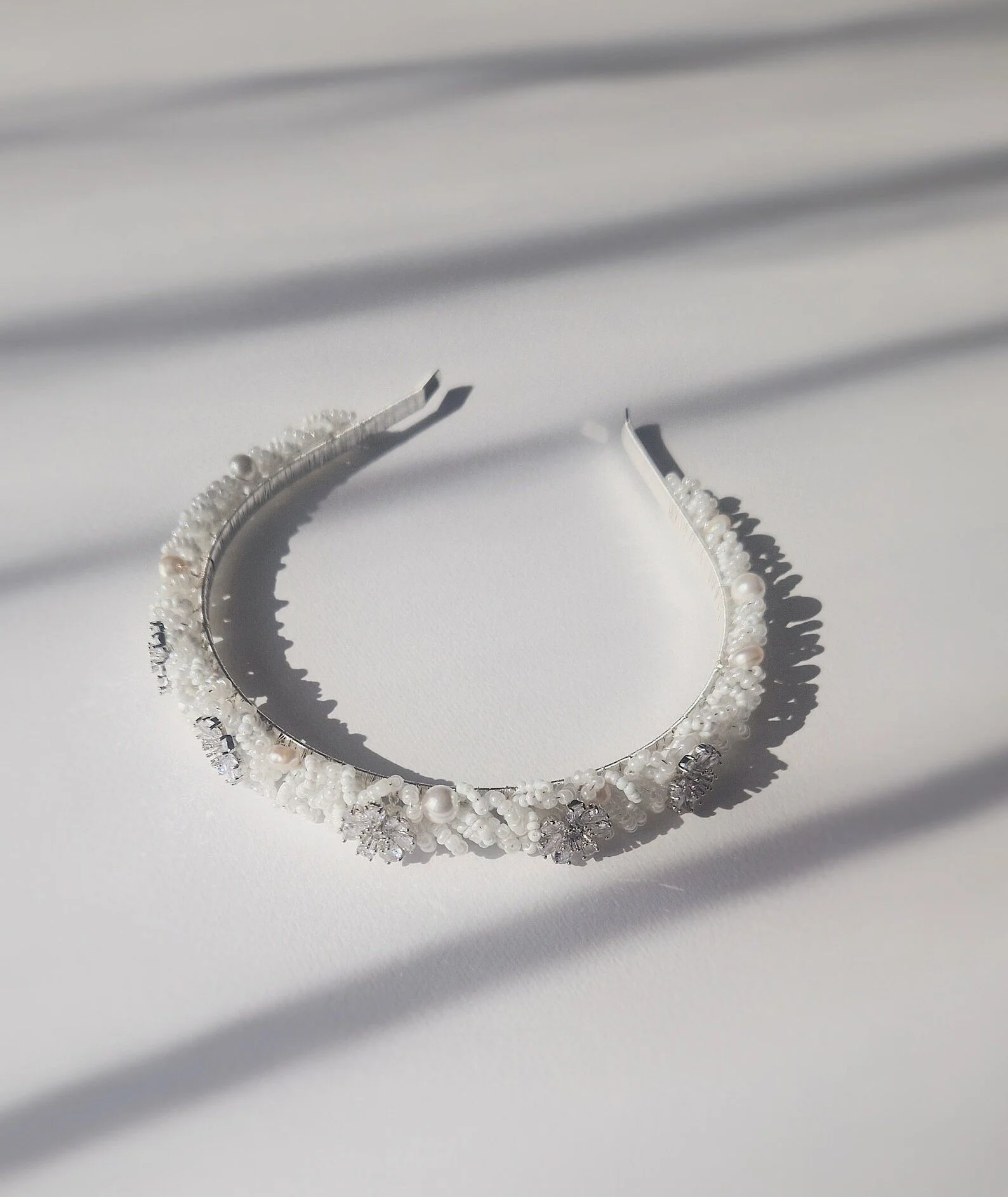 Schmaler weißer Braut-Haarreif, verziert mit kleinen Perlen und dezent funkelnden Kristallblüten, im Sonnenlicht fotografiert.