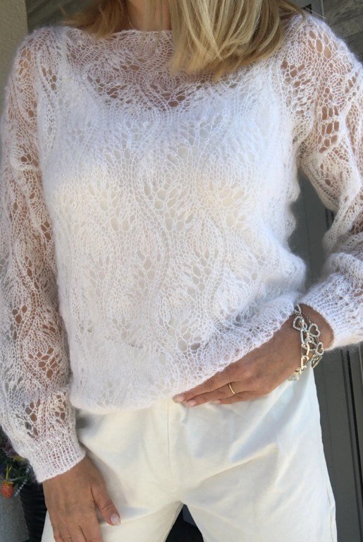 Frau in einem transparent gestrickten weißen Pullover mit Spitzenmuster, kombiniert mit heller Hose und dezentem Goldschmuck.