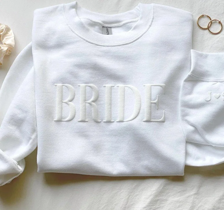Weißer Pullover mit dem Schriftzug „BRIDE“ in 3D-Stickerei, daneben ein Ärmel mit Herzinitialen, zwei Ringe und ein Scrunchie, flach auf Bettwäsche arrangiert.