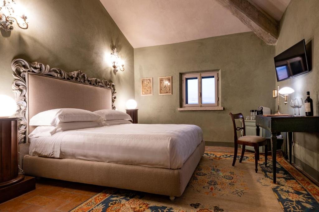 Foto eines gemütlichen Schlafzimmers mit salbeigrünen Wänden, einer schrägen Holzbalkendecke und einem großen Bett mit kunstvoll verziertem Kopfteil auf einem Orientteppich, daneben ein kleiner antiker Holzschreibtisch.