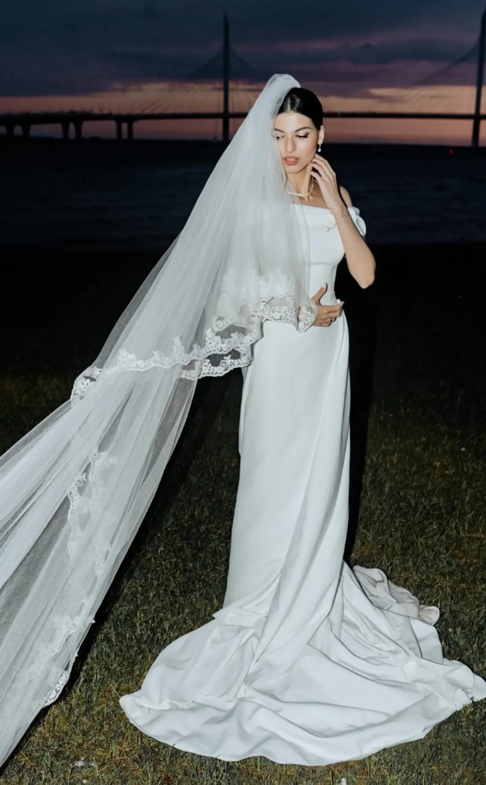 Eine Braut in einem weißen, bodenlangen Brautkleid und mit einem wehenden, verzierten Schleier steht in der Abenddämmerung vor einer Brücke im Hintergrund im Freien.