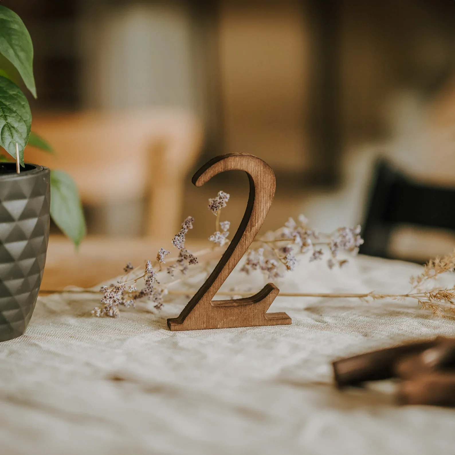 Eine filigrane Tischnummer „2“ aus dunklem Holz, die frei auf einem hellen Leinentischläufer neben dezenten Trockenblumen steht.