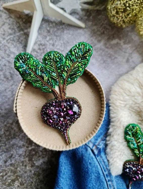 Handgefertigte Brosche in Rote‑Bete‑Form aus Perlen und Kristallen: violette Knolle, grüne Perlenblätter mit Pailletten. Liegt auf runder Pappschachtel neben Jeans. Hochwertiger Broschenschmuck.