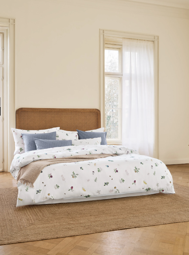 Helles Schlafzimmer mit Parkett und Weiden-Kopfteil. Das Bett hat weiße Bettwäsche mit kleinen floralen Motiven, blaue Kissen und eine braune Decke am Fußende.