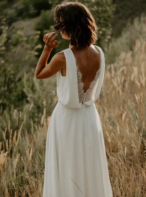 Rückansicht einer Braut in einer herbstlichen Wiese. Das Kleid besticht durch einen tiefen V-Ausschnitt am Rücken, der mit filigraner Spitze unterlegt und einer feinen Knopfleiste verziert ist.
